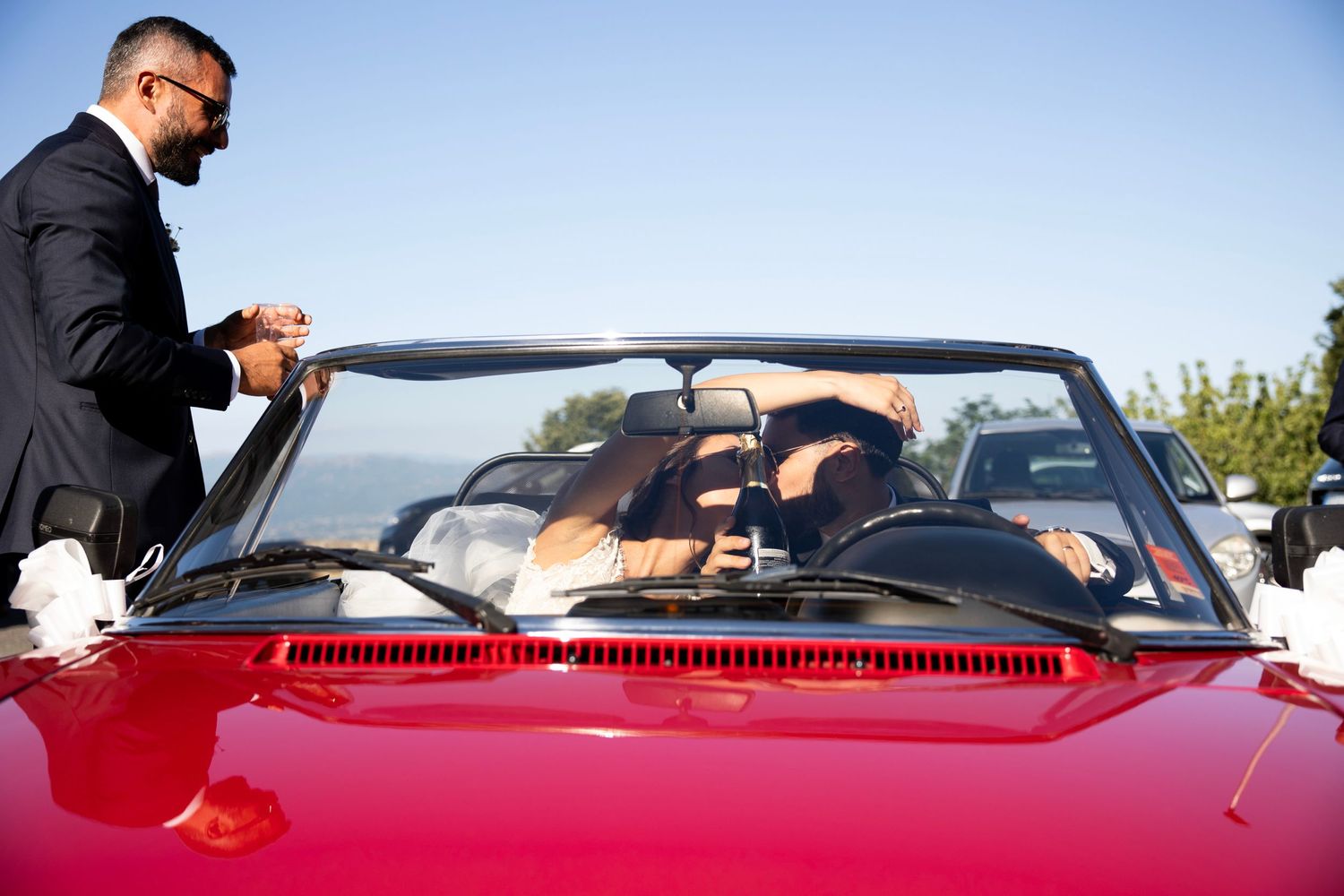 A wedding party gathers around a bright red convertible car under blue skies.