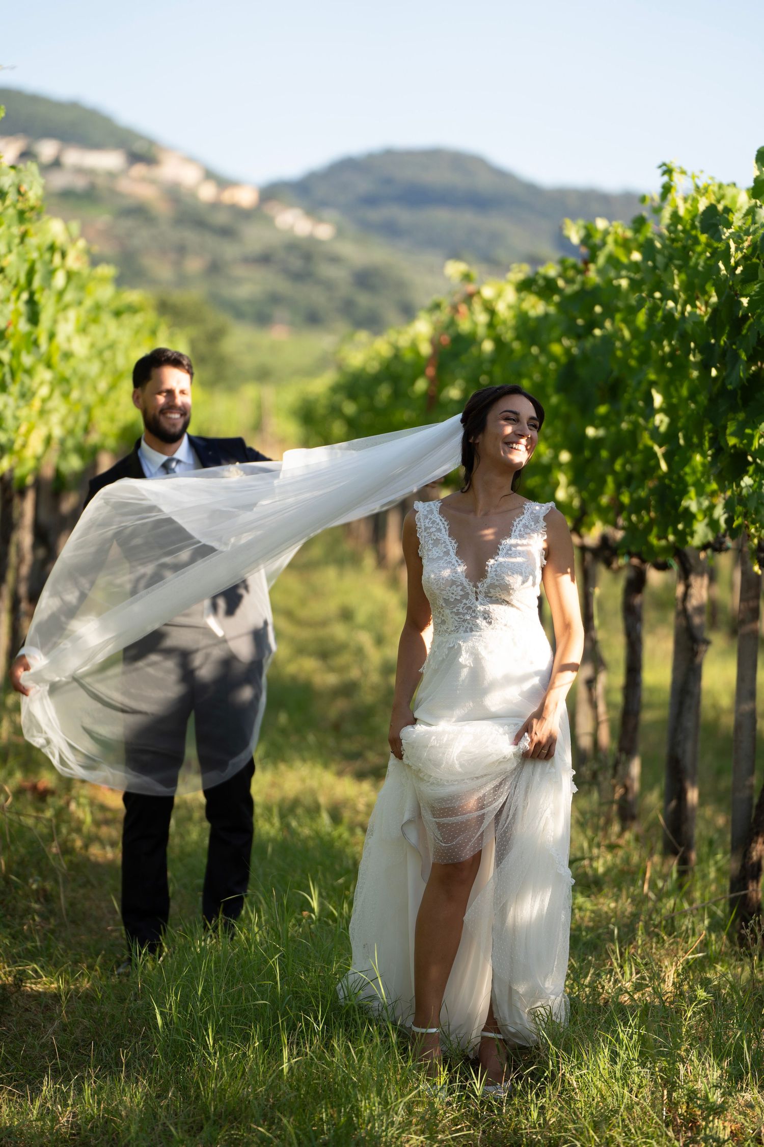 A wedding couple poses playfully in a vineyard as the bride's veil flows in the wind.