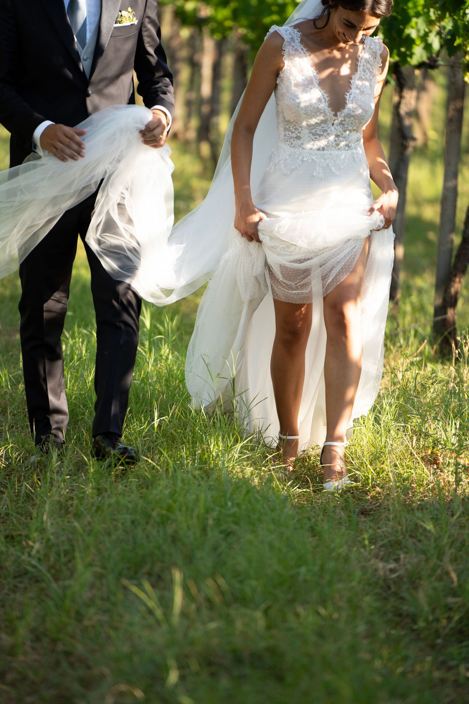Newlyweds holding wedding dress train while walking through grass during outdoor photoshoot.