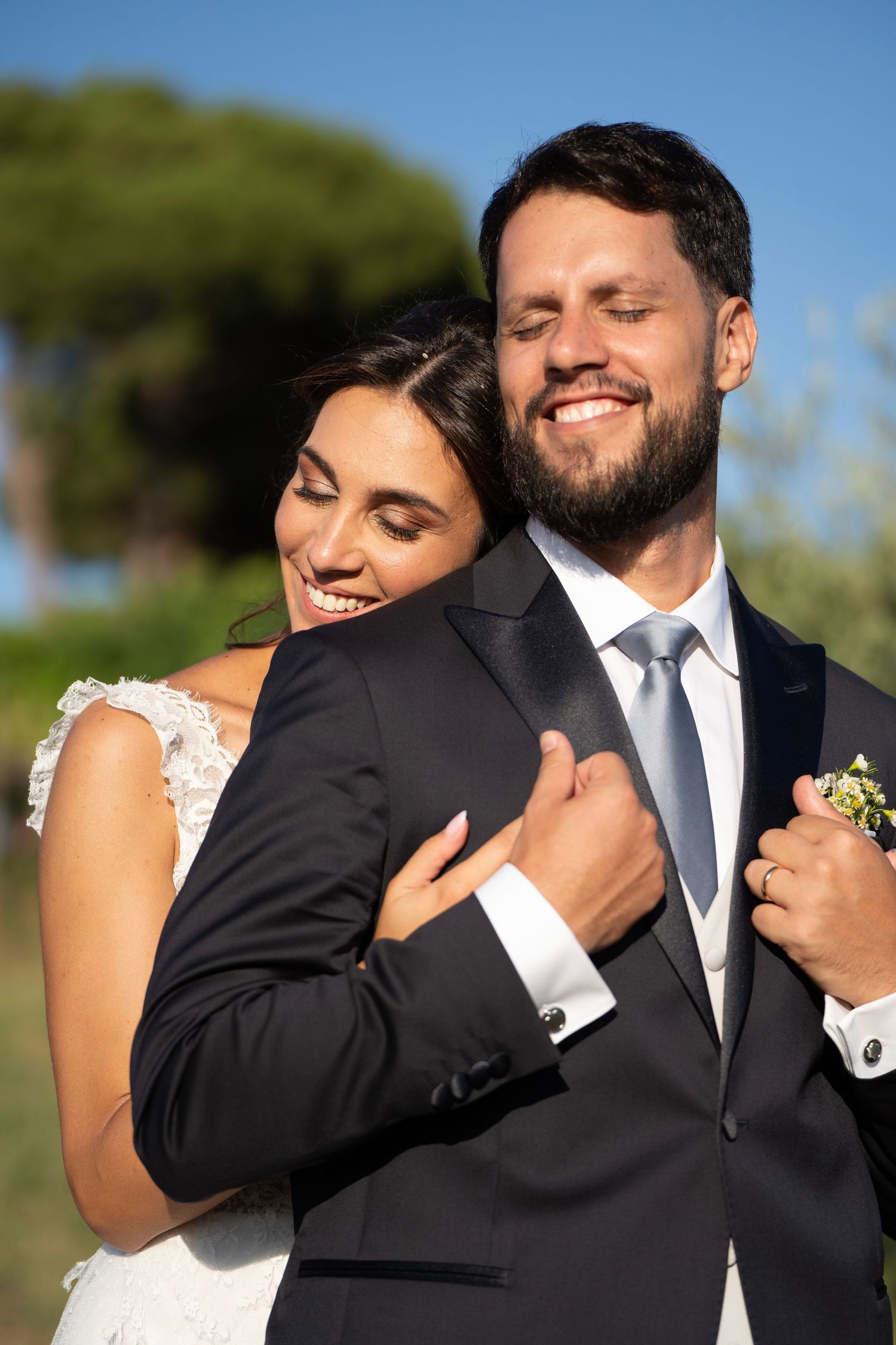 A tender moment between newlyweds in formal attire against a sunny outdoor backdrop.
