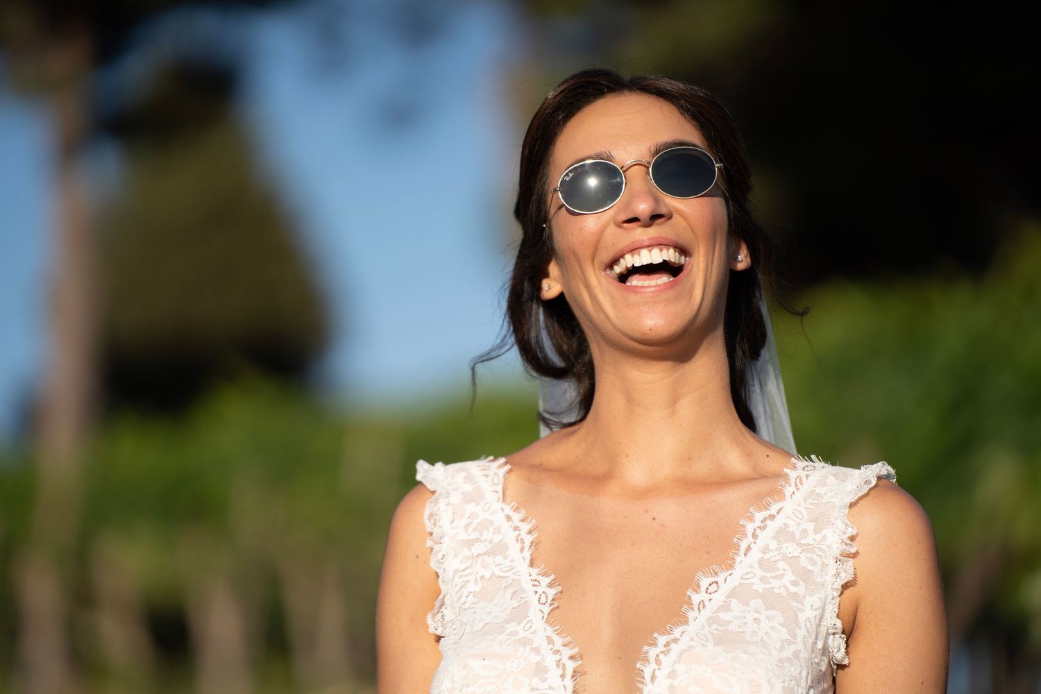 A happy bride wearing sunglasses and a lace wedding dress smiles outdoors.