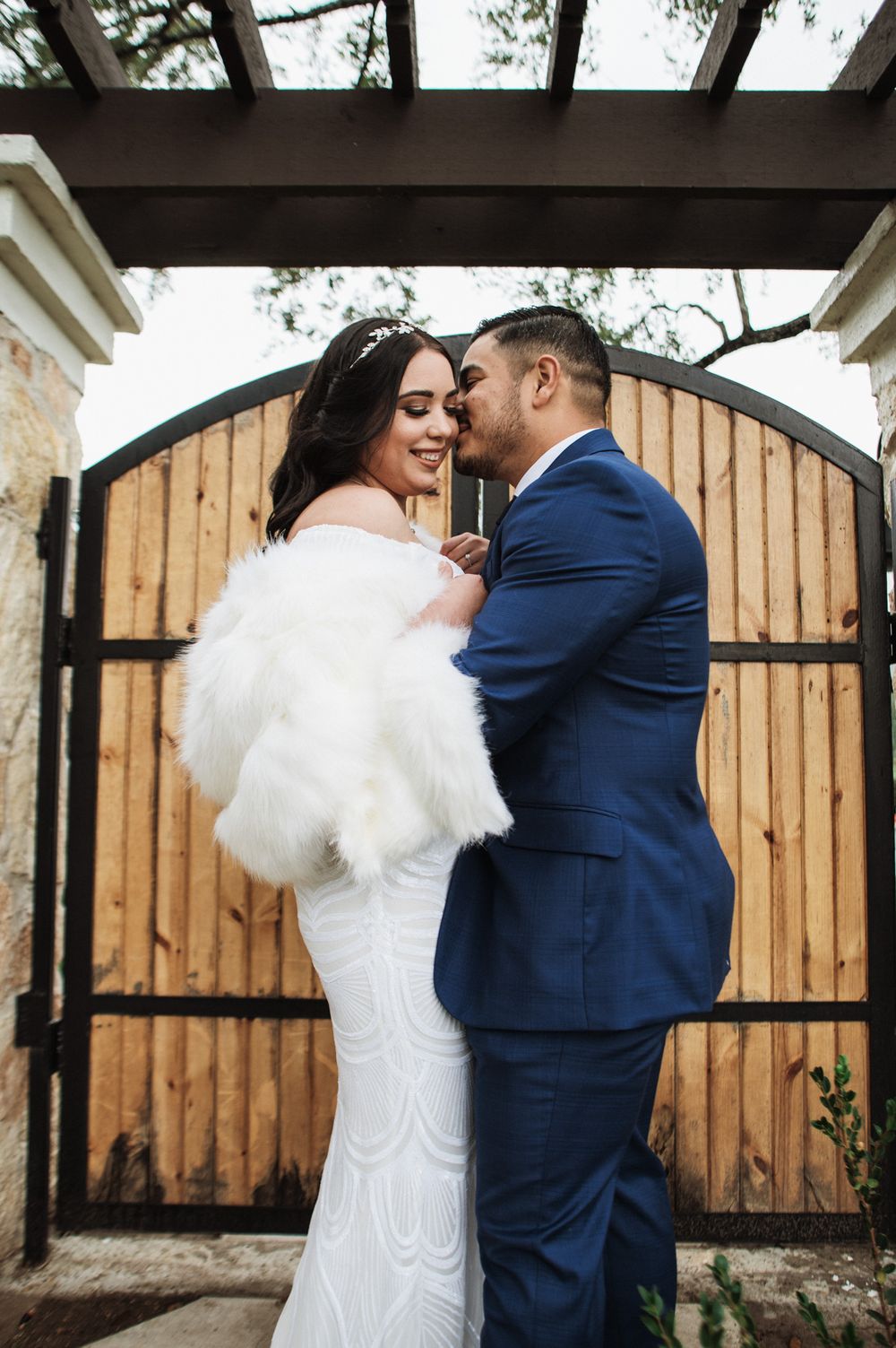 Romantic wedding photo of couple by wooden gate, bride in white dress and groom in navy suit.