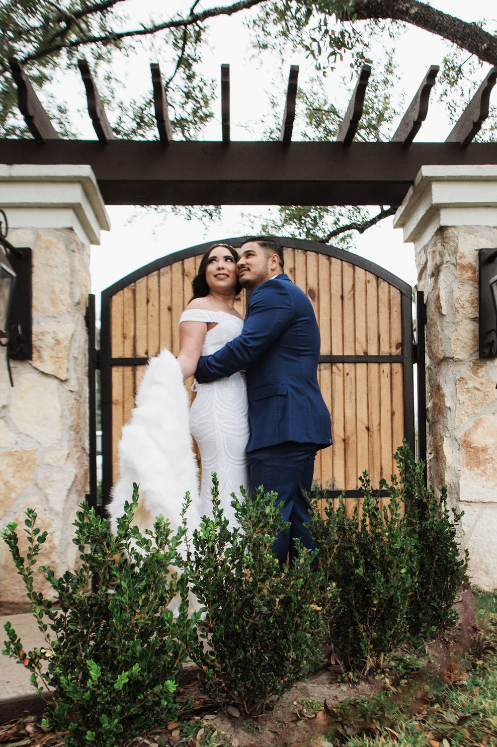 Newlyweds share an intimate moment under a rustic wooden pergola with stone pillars and garden landscaping.