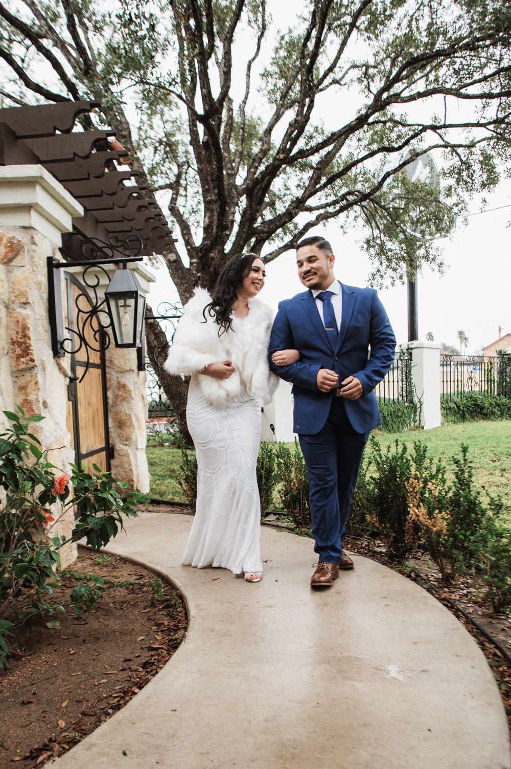 A joyful couple strolls along a curved pathway under trees, with the bride in white and groom in navy.