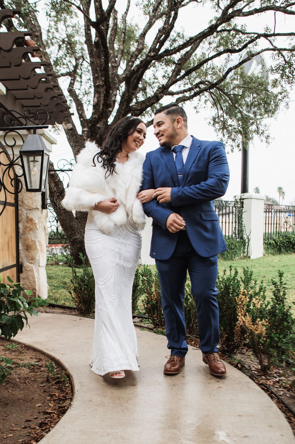 A wedding couple walks together along a garden path, with the bride in a white gown and groom in navy suit.