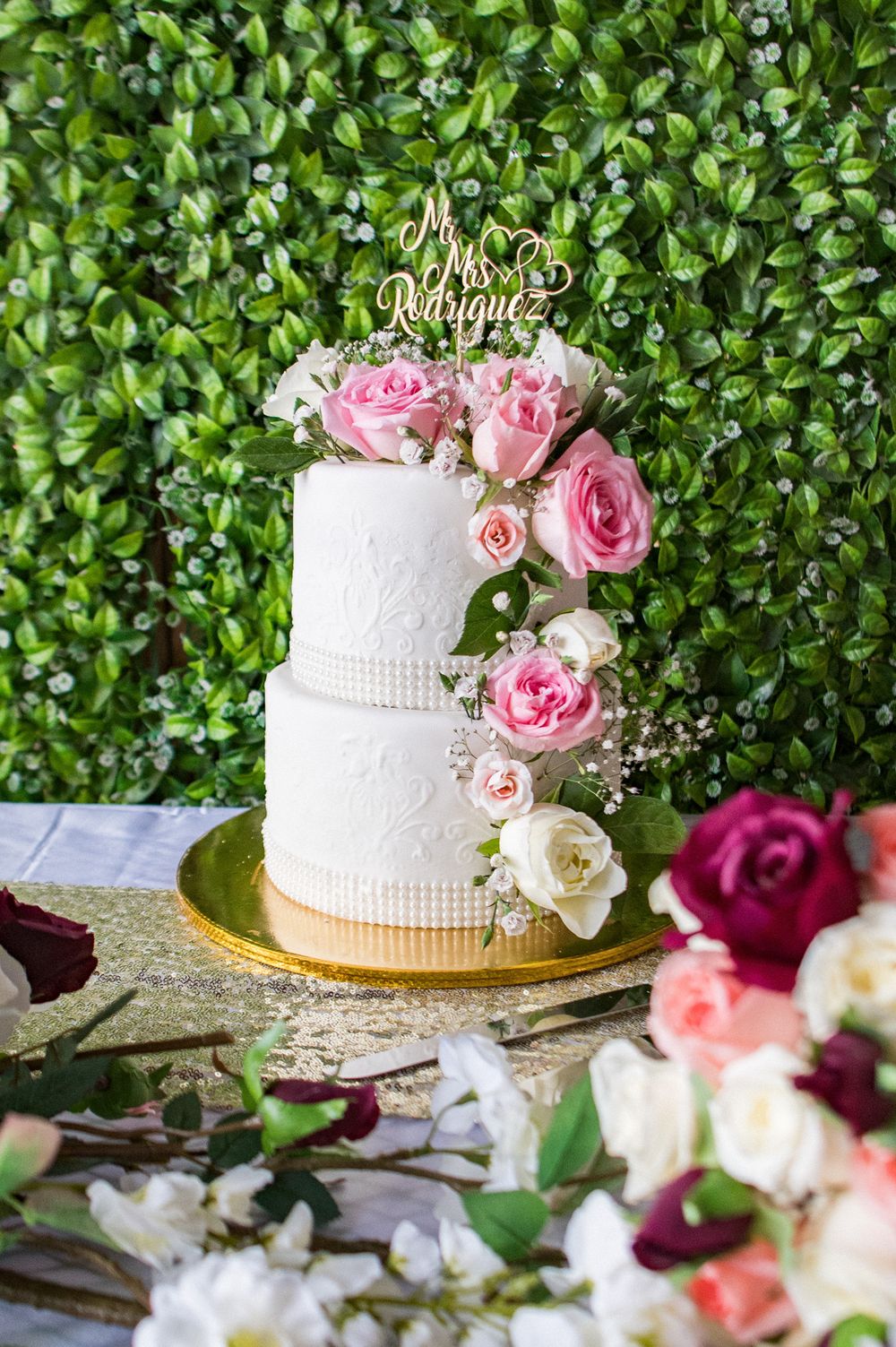 Two-tiered white wedding cake adorned with pink and white roses against a lush green hedge background.