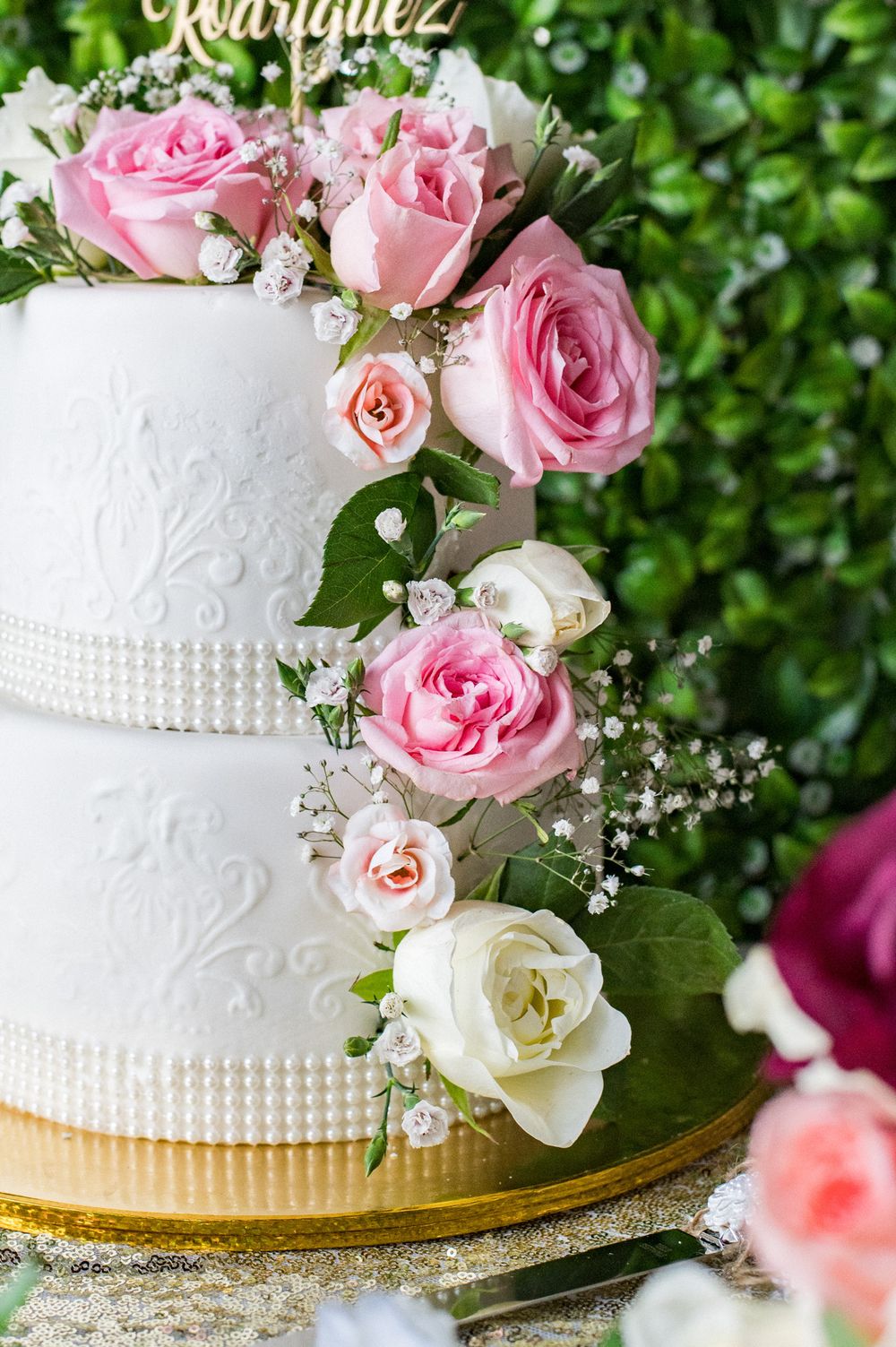Elegant white wedding cake decorated with fresh pink roses and pearled details against a green backdrop.