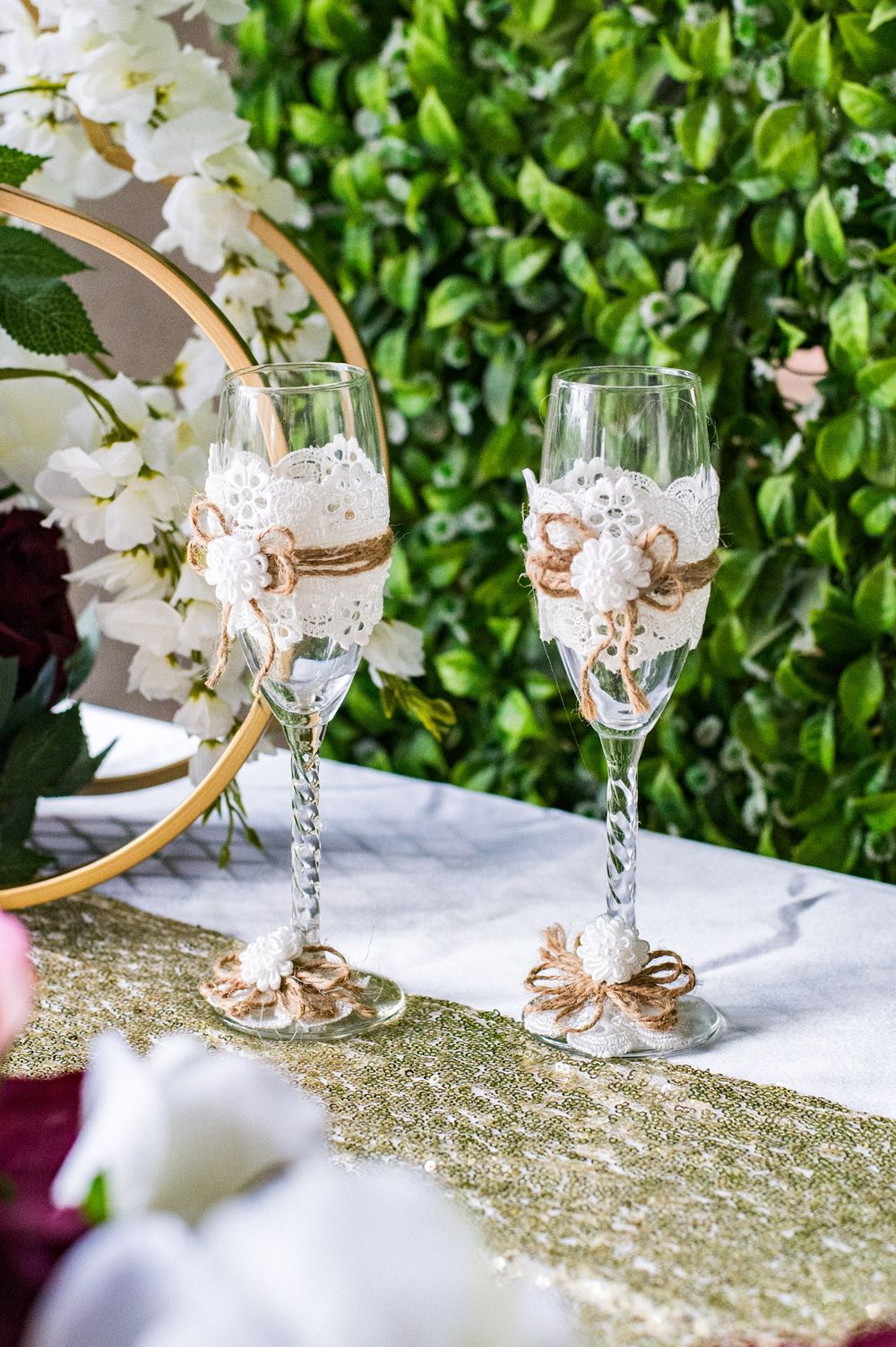 Decorated champagne flutes with floral accents on a table runner with greenery backdrop.