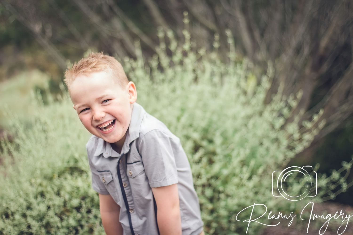 Happy young boy laughing outdoors in a natural garden setting wearing a grey shirt.