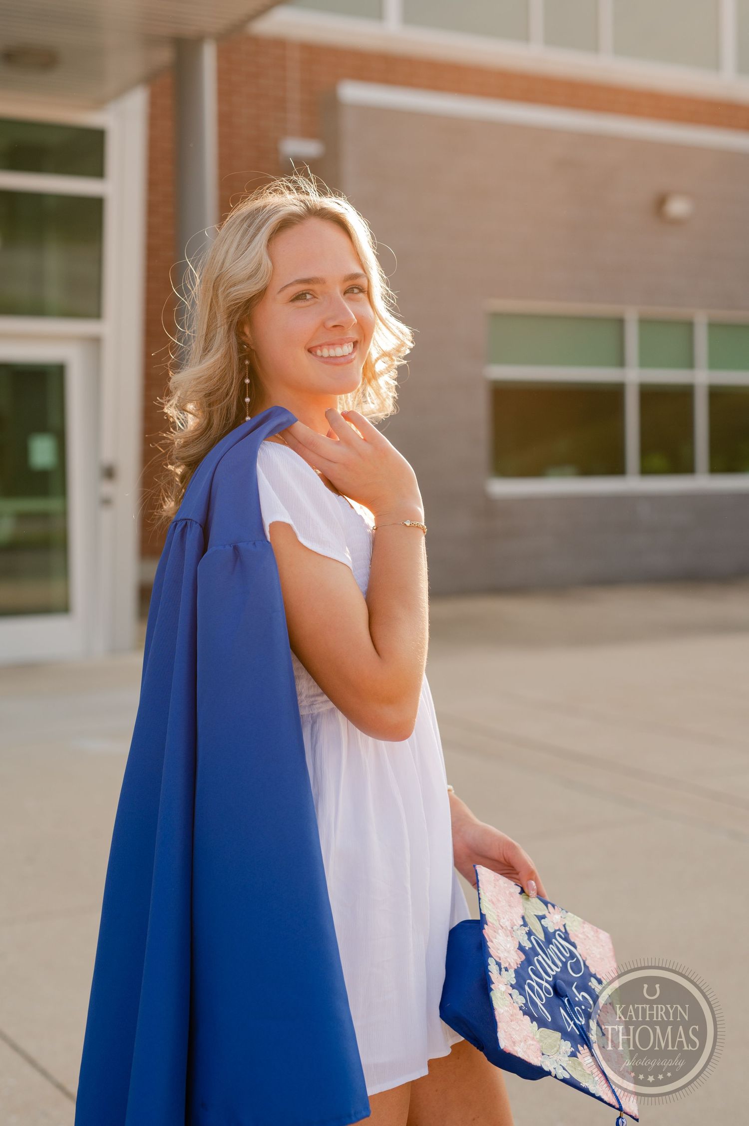 Graduate in white dress and blue gown smiles during sunset photoshoot outside school.