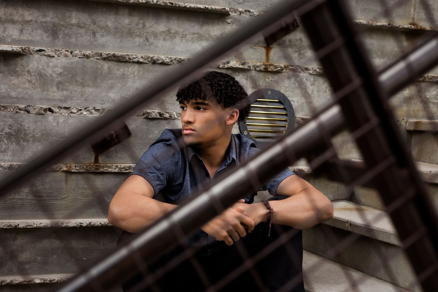 Cool senior portrait of graduate boy where the camera view is peering at the senior through the fence during his graduation photoshoot.