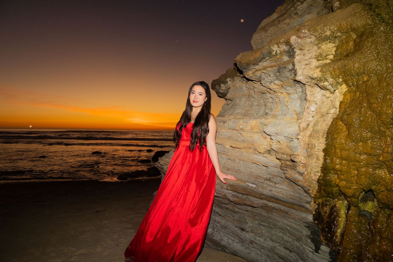 A high school senior in red gown poses against rocky cliff during dramatic beach sunset.