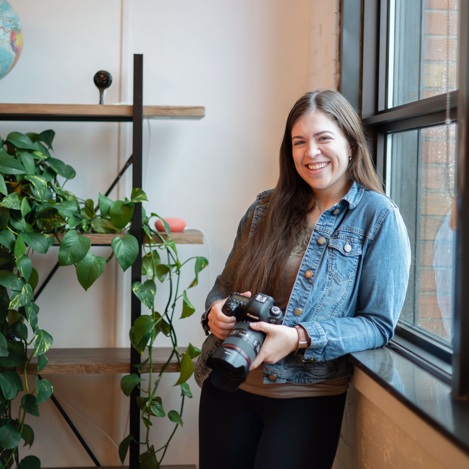 A photographer holding her camera leaning against a window next to a shelf with plants