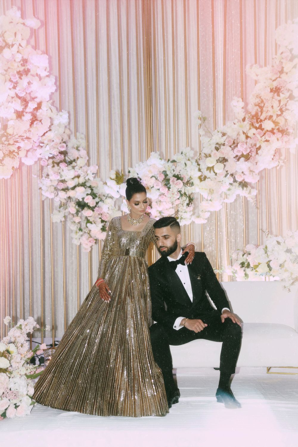 Glamorous couple in gold gown and black tuxedo pose against floral backdrop.