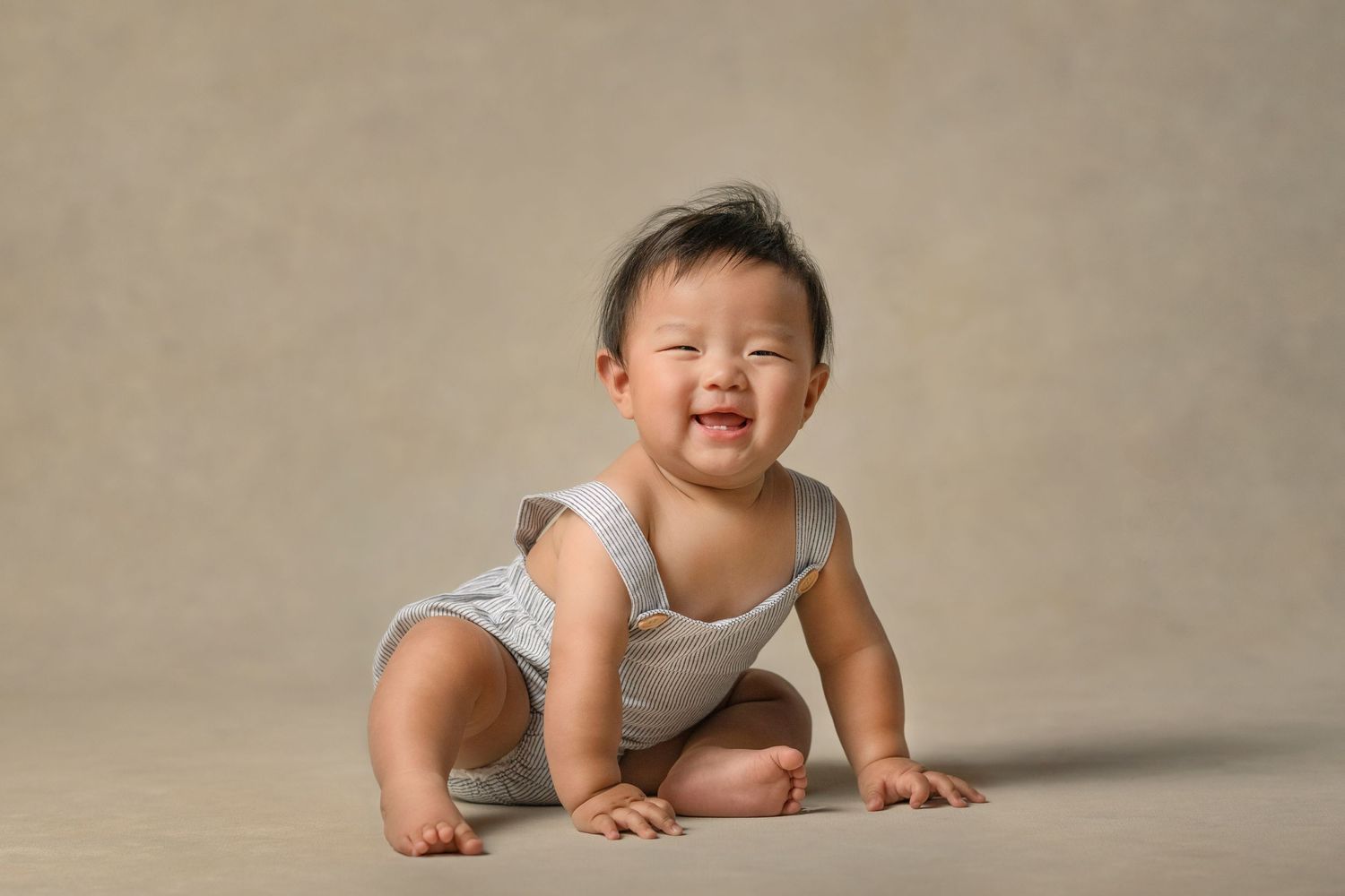 Adorable smiling six month old baby in light blue romper sits on a classic beige canvas portrait backdrop.