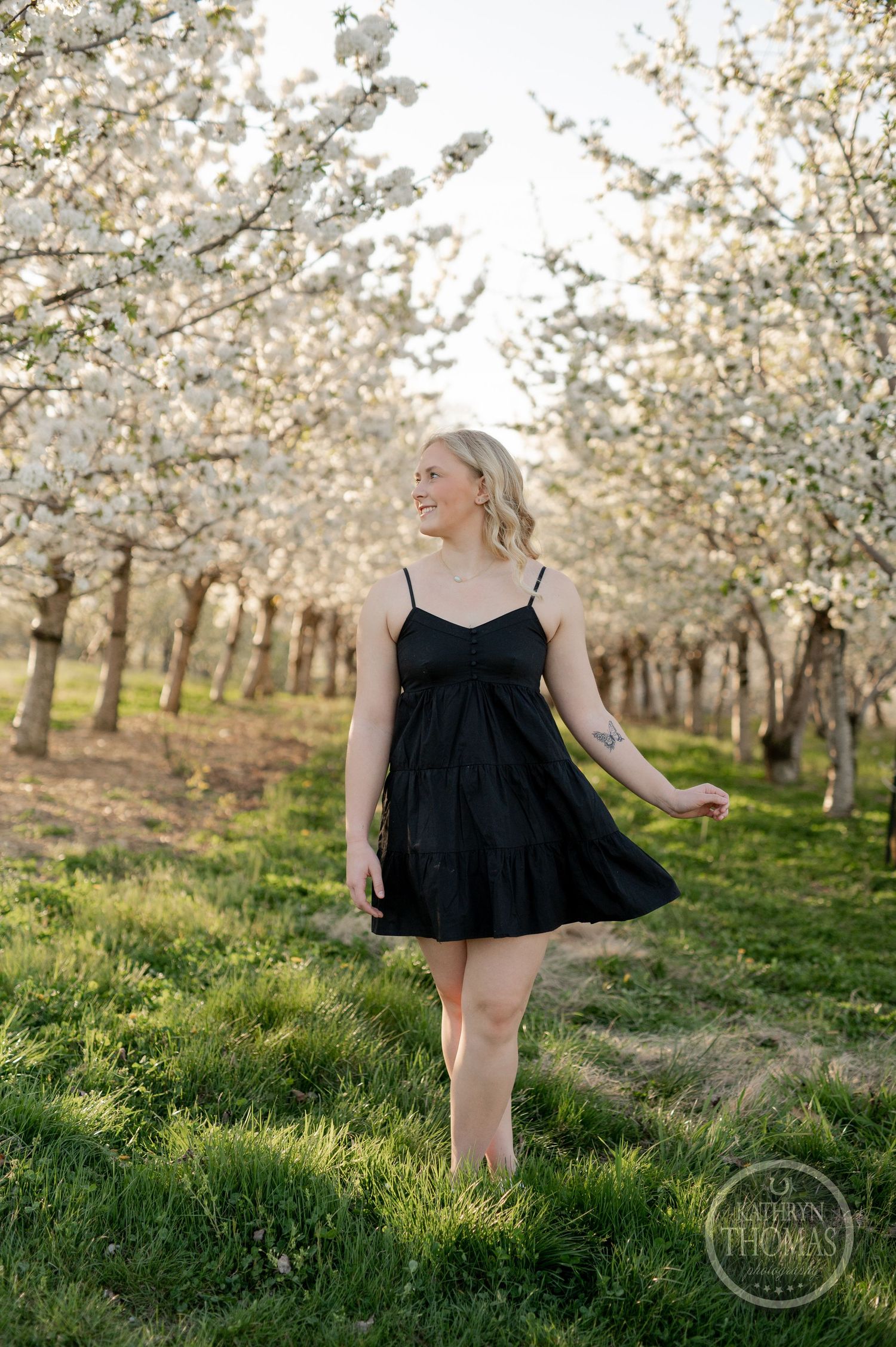 Figure in black dress stands gracefully among blooming cherry trees in an orchard setting.