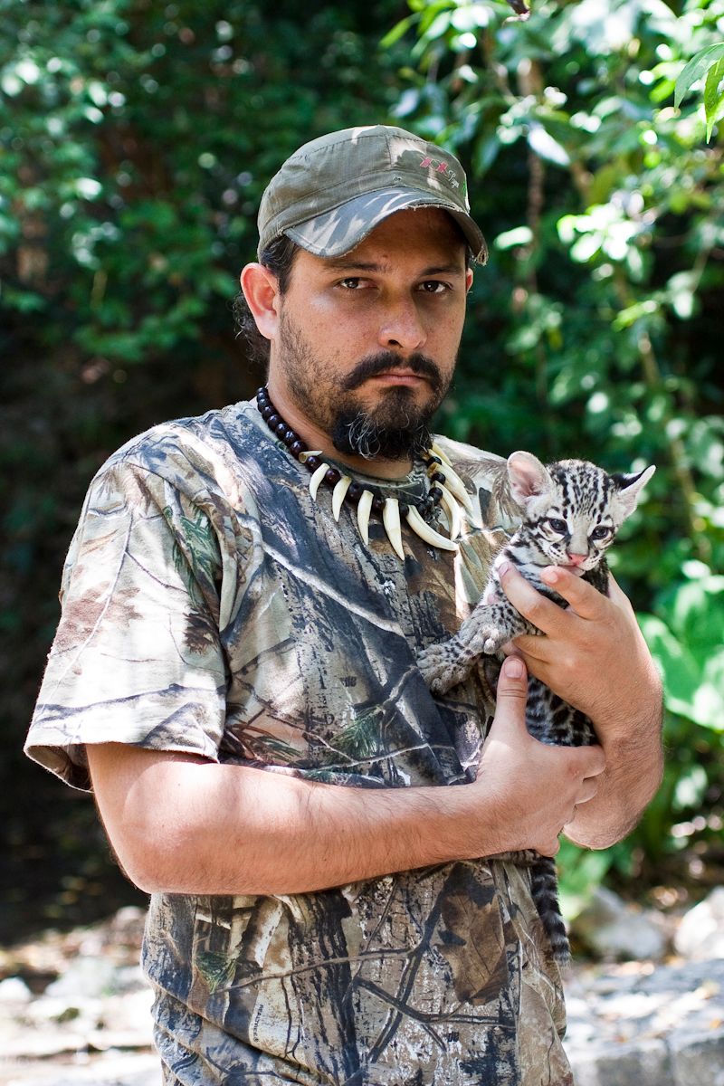 Person in camouflage clothing holds a small spotted wild cat in an outdoor setting.