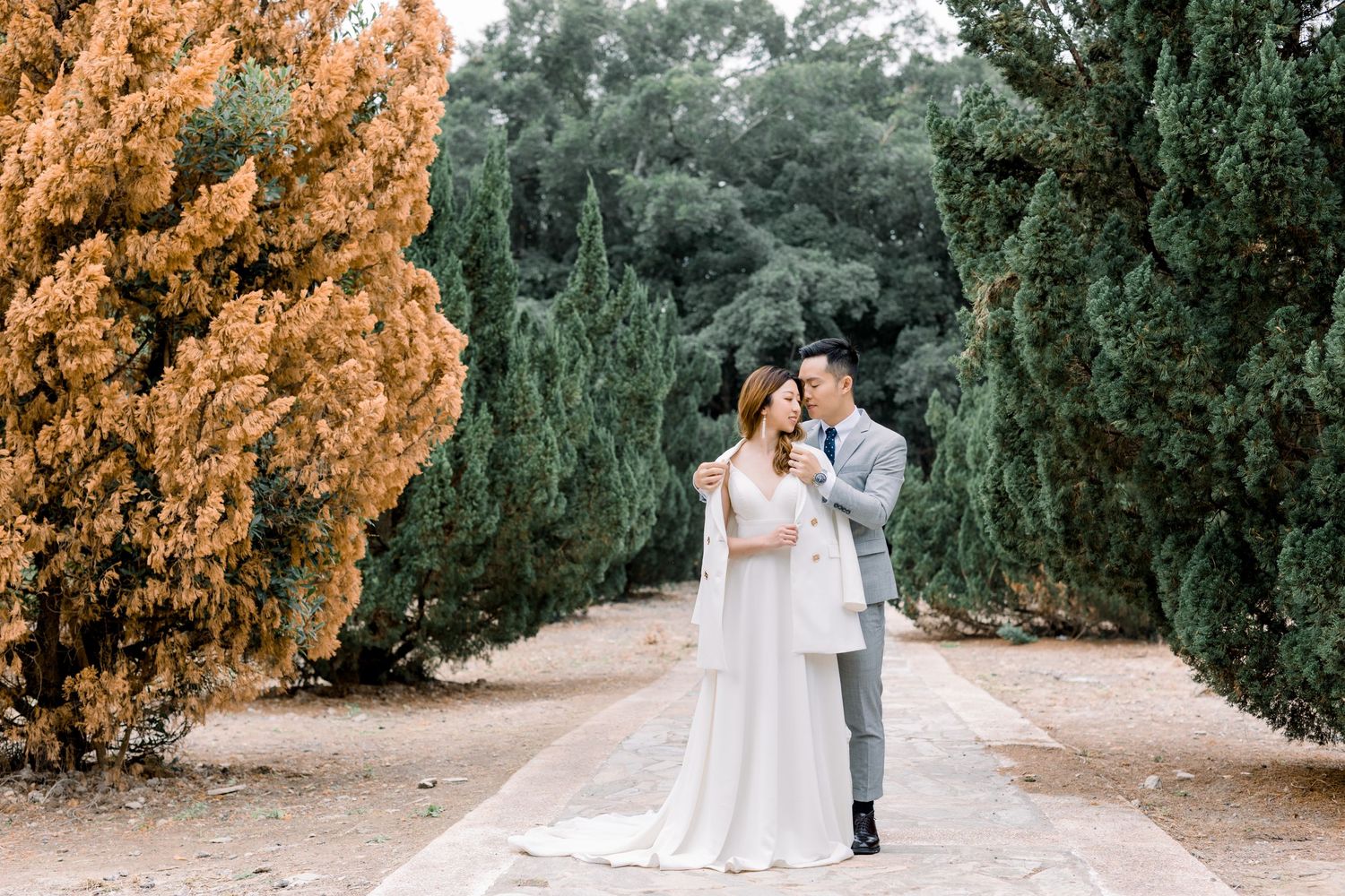 Wedding couple poses on a garden path lined with evergreen trees and golden foliage.