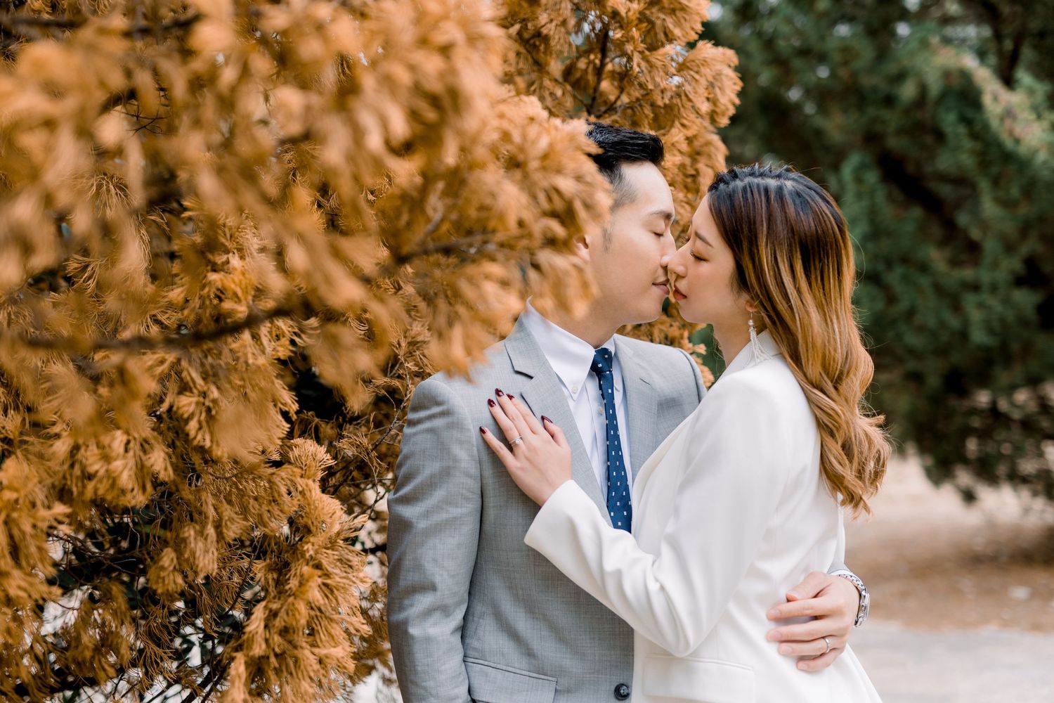 A tender embrace between a couple in formal wear beside golden autumn trees.