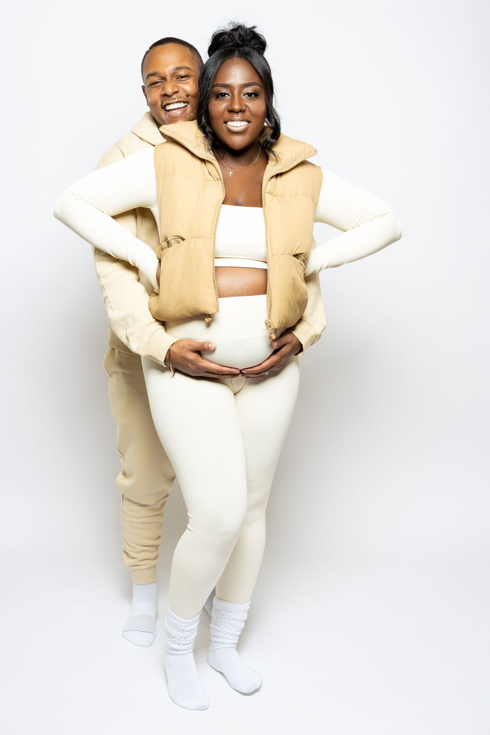 Expectant couple in coordinated cream outfits share a joyful moment during maternity photoshoot.