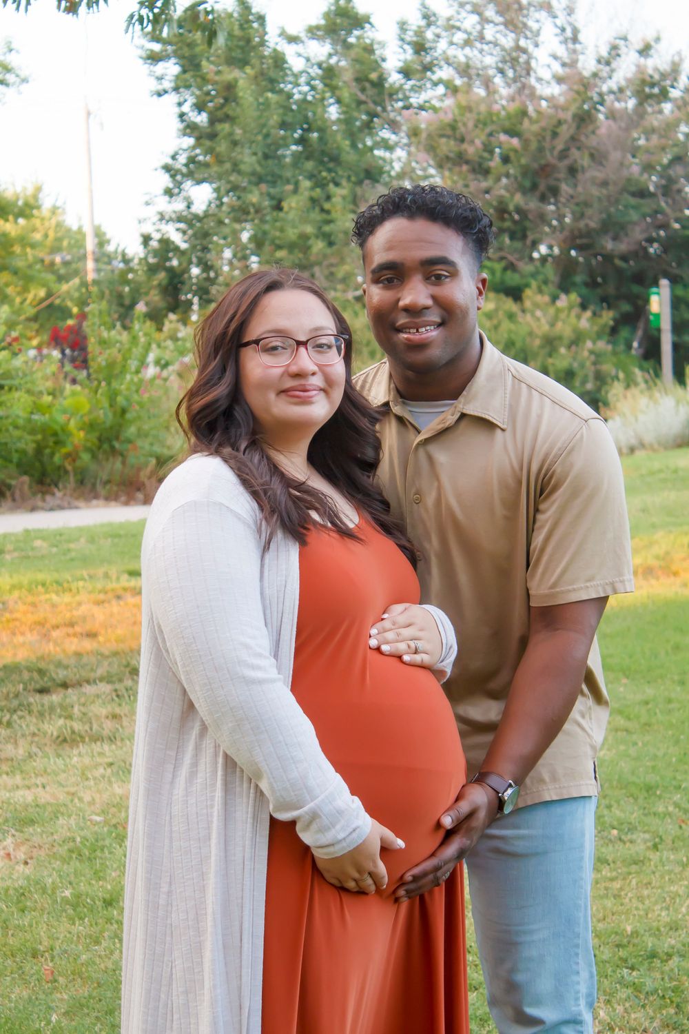 An expectant couple poses together in a garden setting during golden hour.
