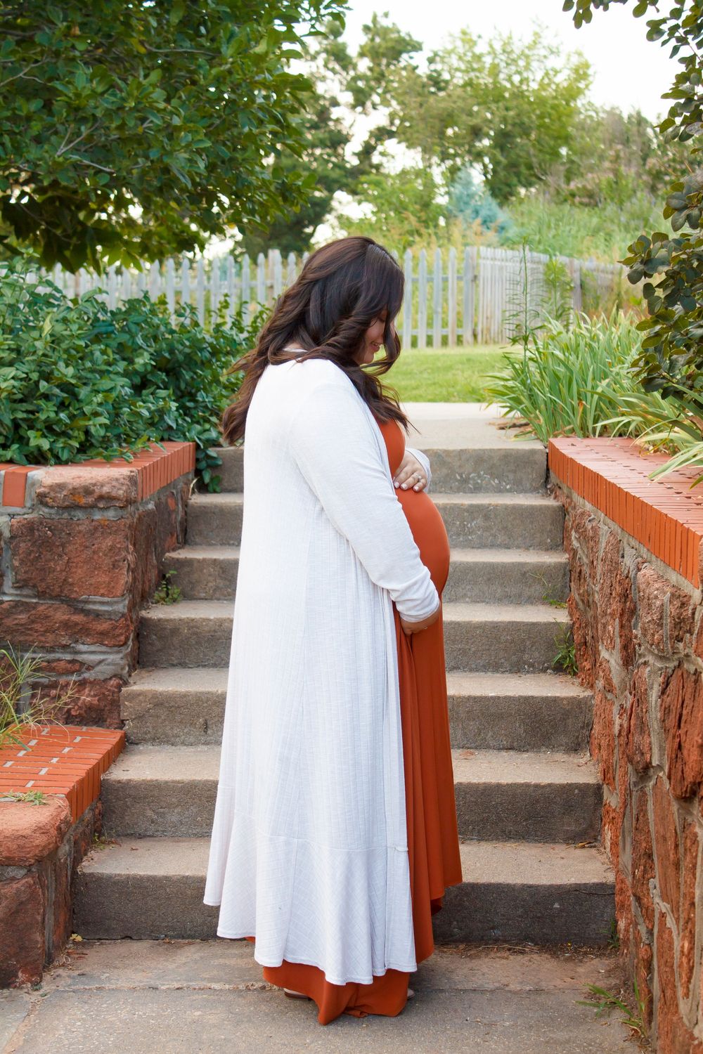 A side view of someone in a white cardigan and rust-colored dress standing on brick steps in a garden.