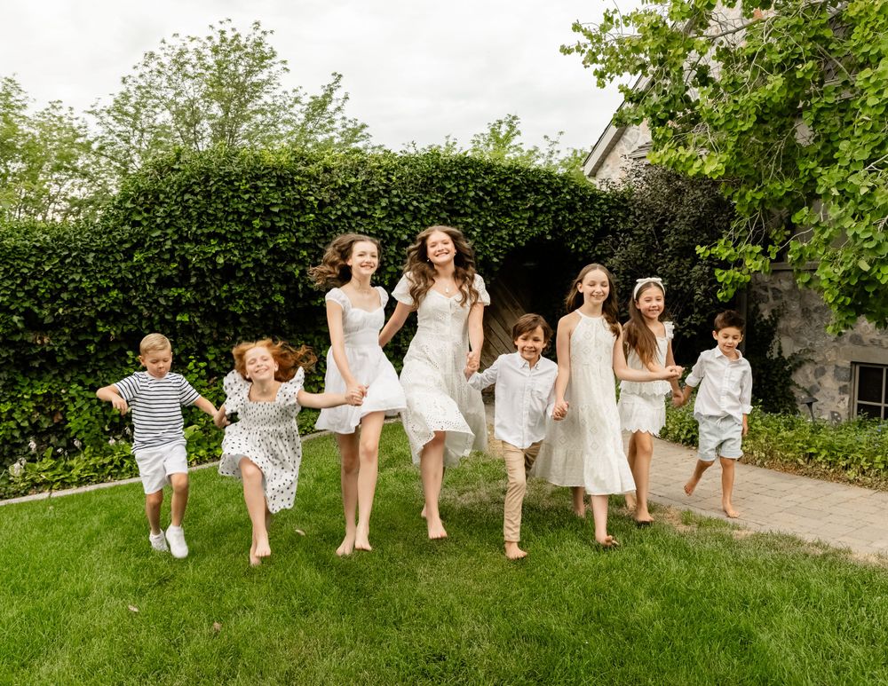Children and young people run joyfully across a green lawn in coordinated white summer outfits.