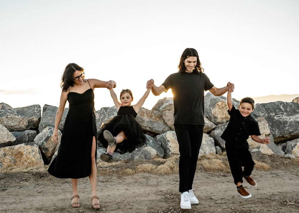 A group in black outfits holds hands and jumps together on rocky terrain at sunset.