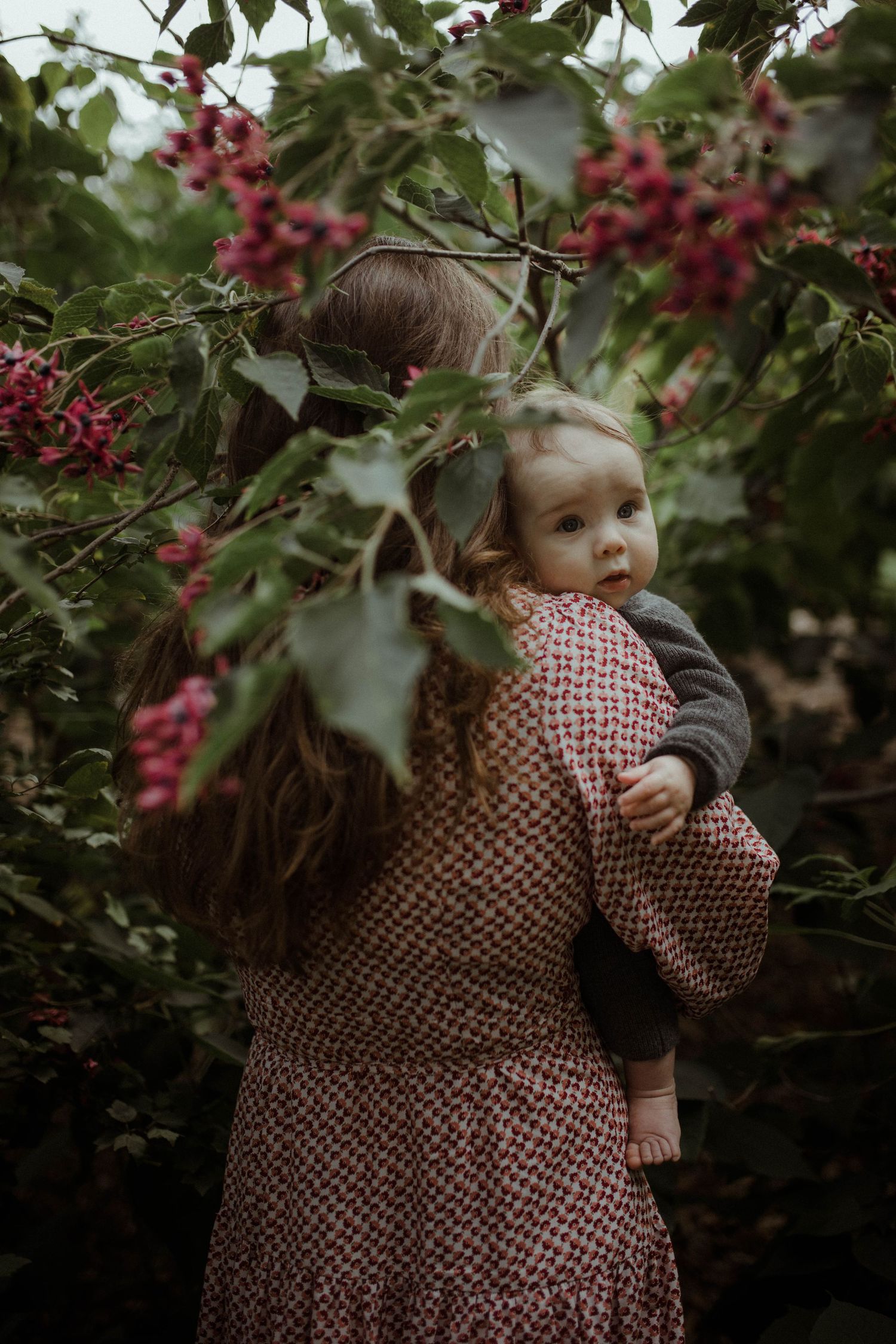 Mother carrying a wide-eyed baby through a lush garden with vibrant pink berry-laden branches overhead.
