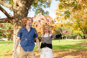 Couple playing with falling autumn leaves in park during golden hour.
