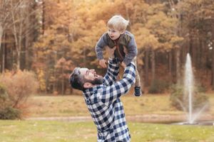 Parent lifting child joyfully in autumn scene with fountain in background.