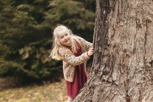 A young child peeks playfully around a large tree trunk in an outdoor setting.