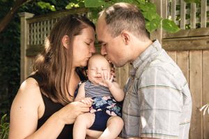 Parents share an intimate moment with their baby outdoors against a wooden fence backdrop.