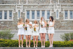 Five women in matching white dresses pose together against stone university building.