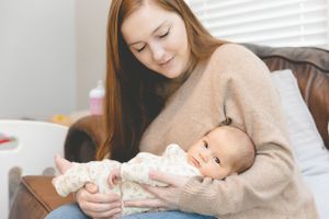 Tender moment as a caregiver in beige sweater bottle feeds a small infant on a brown leather couch.