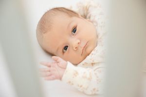 Close up portrait of a baby with light hair and bright eyes looking through crib slats.
