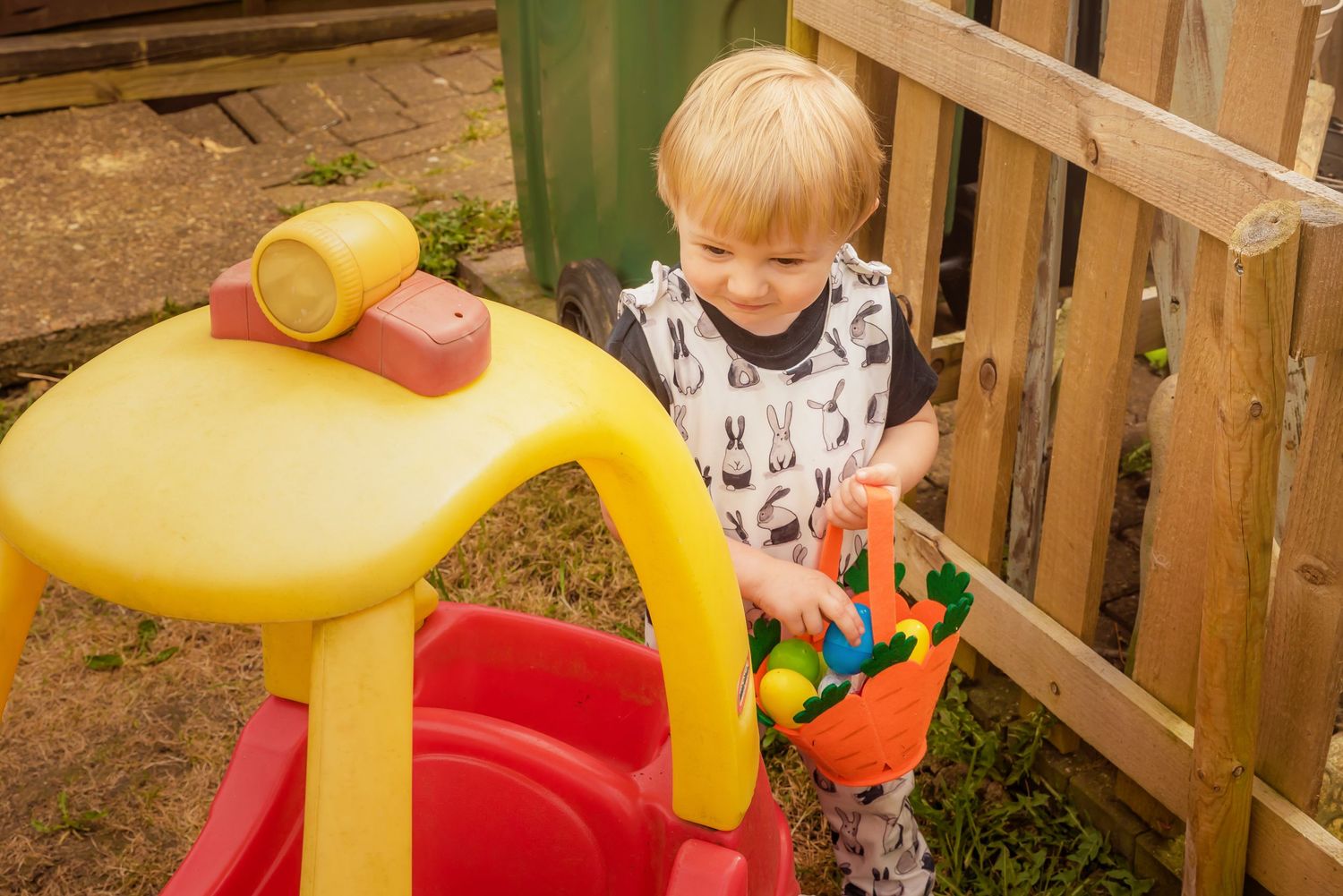 A child plays with toys on a yellow plastic play table near a wooden fence.