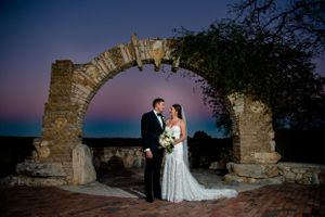Wedding couple posing under stone archway at twilight near ocean.