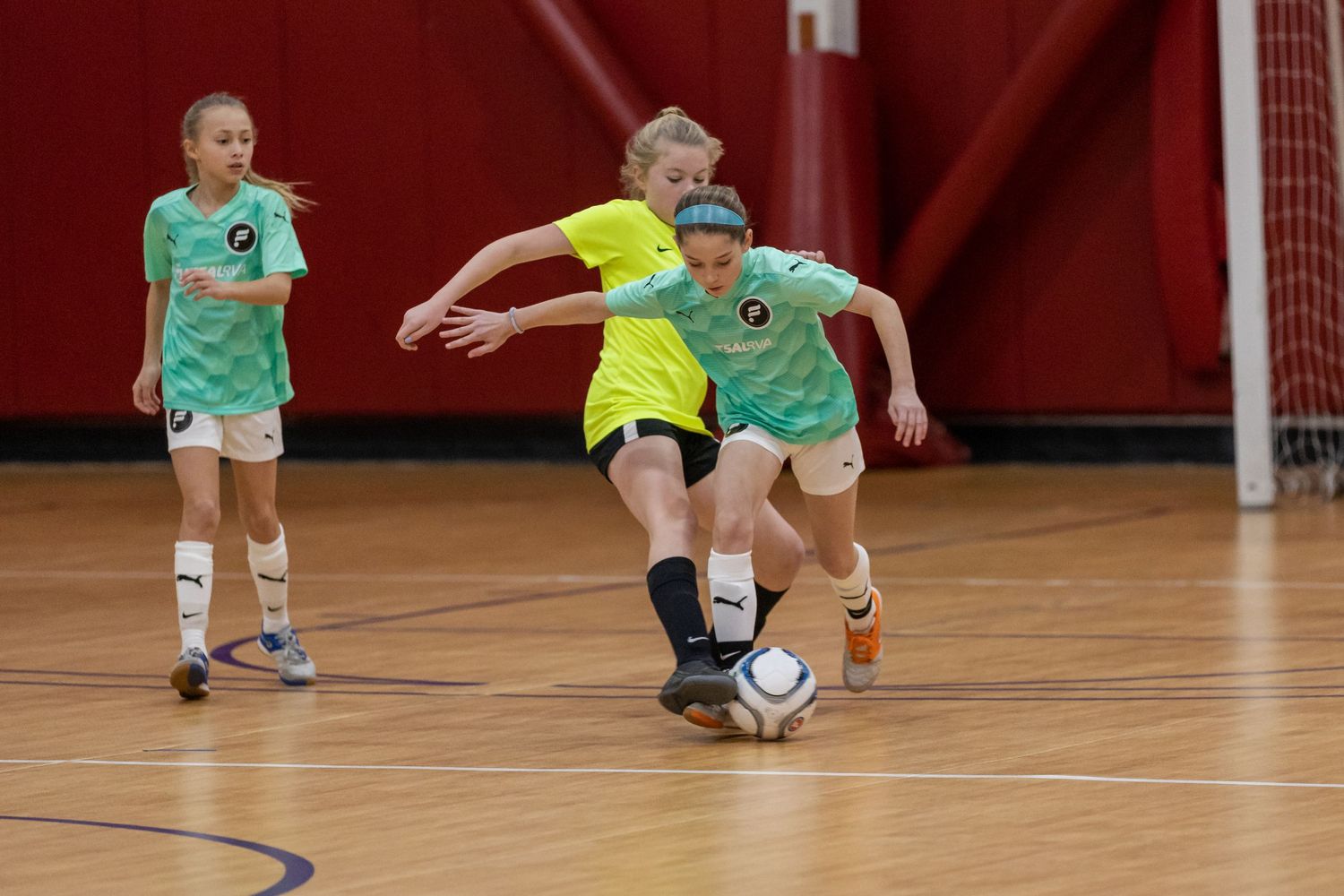 Children playing indoor soccer in colorful uniforms on gymnasium court.