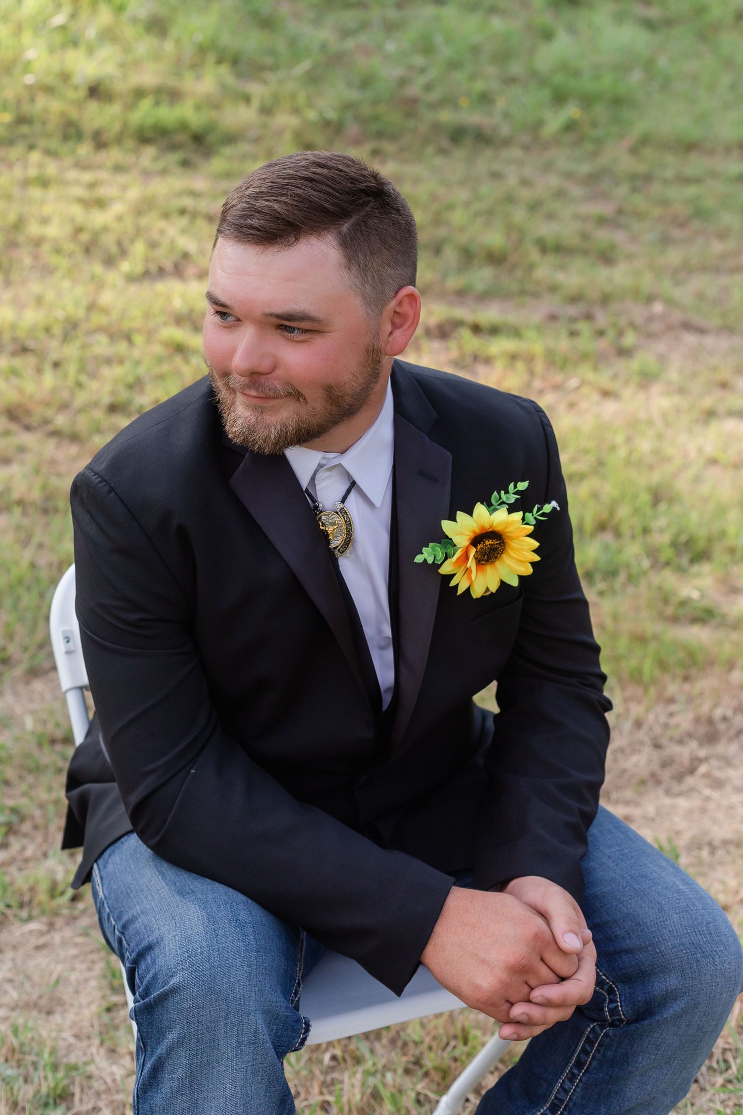 Groomsman in dark suit with sunflower boutonniere sits casually outdoors.