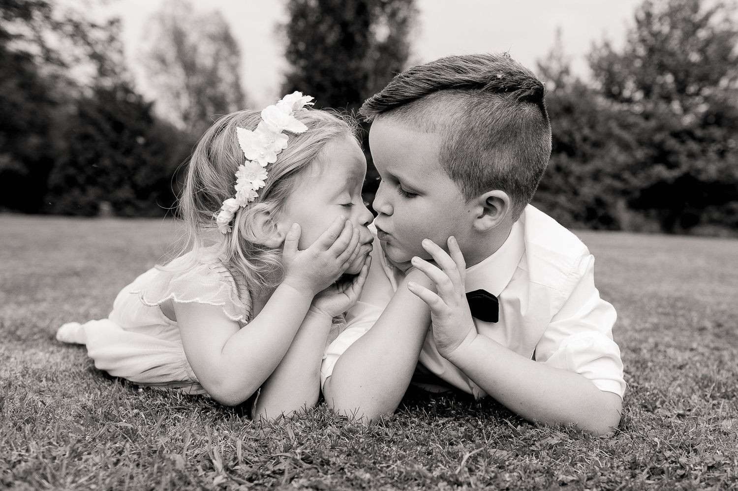 Two young children in formal wear lying in grass whispering secrets.