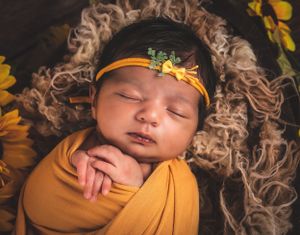 Newborn wearing orange headband wrapped in yellow fabric on textured brown backdrop with sunflowers.