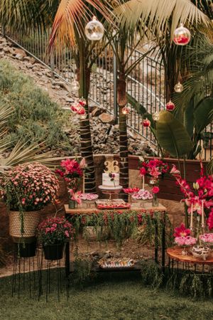 Tropical party setup with pink flowers and hanging red ornaments under palm trees.