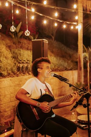 Musician performs on acoustic guitar under warm string lights in an outdoor evening setting.