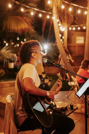 Guitarist singing into microphone during nighttime performance under string lights.