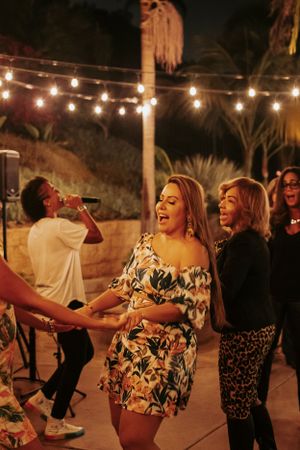 Friends dancing and laughing at an outdoor party under string lights with palm trees in the background.