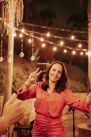Woman in coral outfit making peace sign while dancing under string lights at outdoor party.