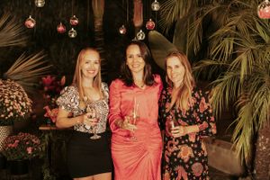 Three friends posing with drinks under hanging lights at tropical outdoor party.