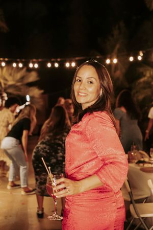 Woman in coral pink dress smiles at outdoor evening party with string lights and tropical decor.