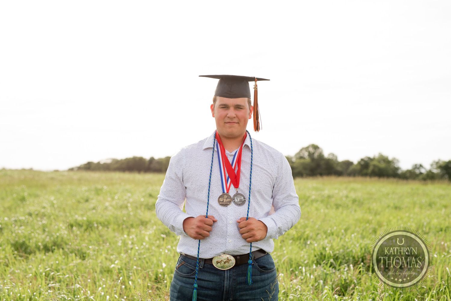 Graduate in graduation cap with medals stands in grassy field at sunset.